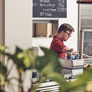 Eine Person steht an der Theke und arbeitet. Es sind Servietten und Besteckbehälter zur Selbstbedienung sichtbar.