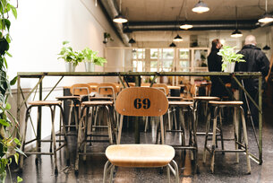 Blick in das Restaurant. Im Vordergrund ein Holzstuhl mit der Nummer "69" auf der Rückenlehne eingebrannt. Im Hintergrund ein Bistrotisch, weiter weg Menschen. An der Decke Rohre und Lampen.