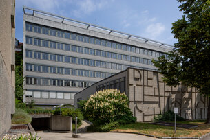 Fassade eines sechs-stöckigen Hauses. Davor ein kleinerer Betonbau und Bäume. 